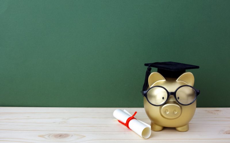 Gfold piggy bank with a grad cap and diploma in front of green chalkboard background. Education scholarship